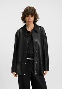 Black leather jacket with a large collar, zip and snap closures, wide sleeves, and a loose fit; soft texture and shiny finish.