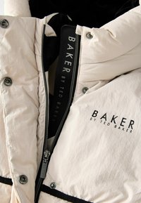 Svijetlo bež jakna s preklopom i ukrasnim šavovima koja ima crni patentni zatvarač, dugmad na preklop i brendiranu oznaku s natpisom "BAKER BY TED BAKER" u podebljanim slovima.