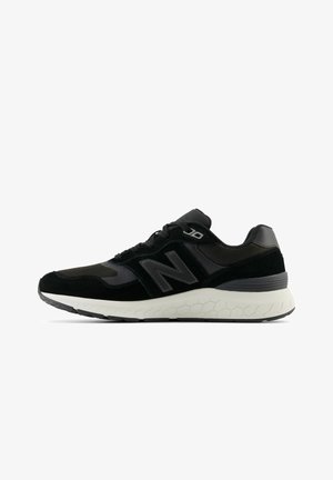 Sneaker nera caratterizzata da un mix di materiali in suede e mesh, con una suola bianca a motivo esagonale e un prominente logo "N". Gli accenti rifiniti sono di un grigio scuro.