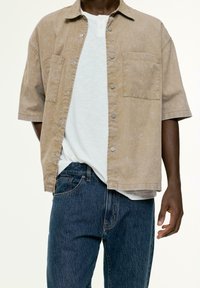 Chemise courte à manches tan avec une texture délavée, deux poches poitrine et des boutons gris, superposée sur un t-shirt gris clair et un jean en denim foncé.