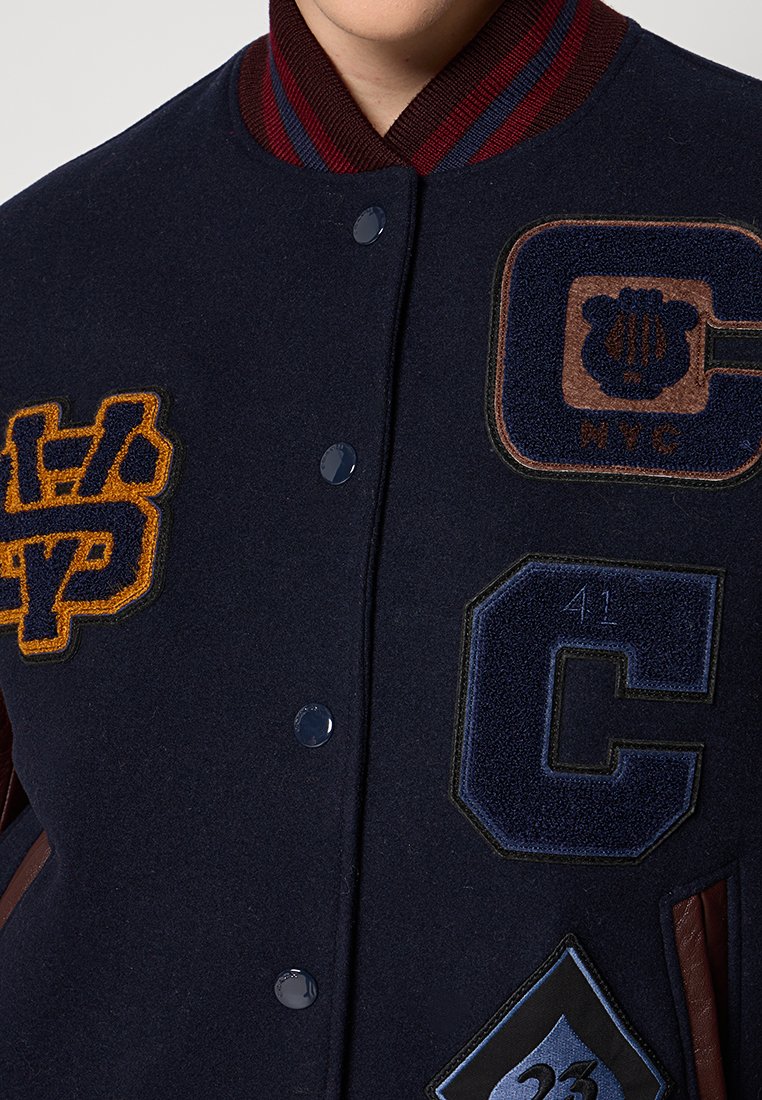 Giacca varsity di lana navy con colletto a coste color bordeaux, numerosi ricami in oro, blu e marrone, e chiusura a bottoni a pressione.