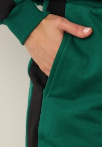 Gröna bomullsjoggers med en svart sidopanel, där en hand syns i fickan. Den ribbade mudden är synlig. Mjuk textur, avslappnad design.