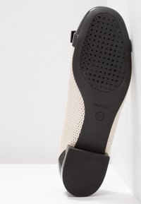 Chaussure beige perforée avec semelle en caoutchouc noir et petit talon, inclinée contre un coin blanc, montrant des trous de ventilation sur la semelle.