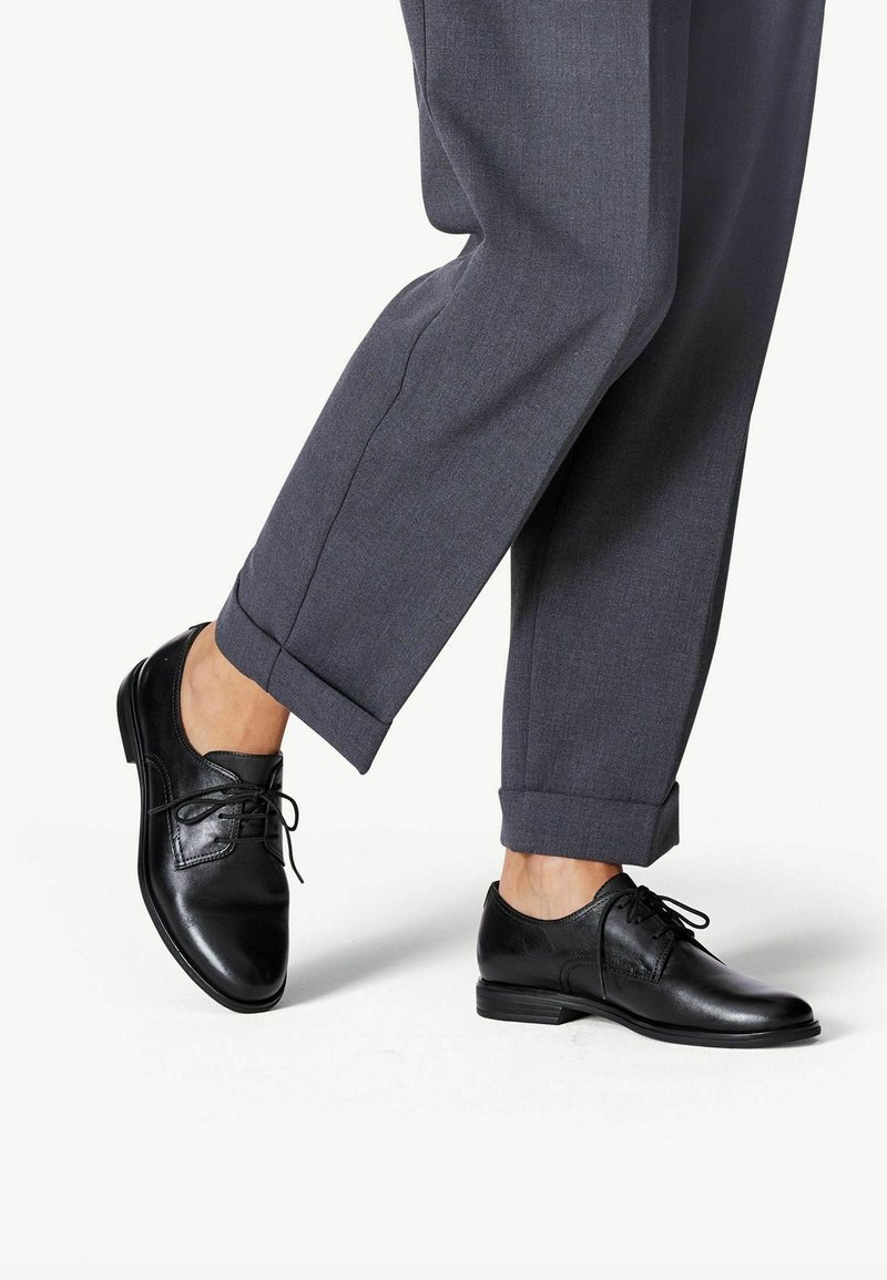 Chaussures Oxford en cuir noir avec lacets, portées avec un pantalon gris retroussé, présentant une texture lisse et une conception à bout arrondi.
