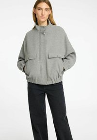 Grijze fleecejas met hoge kraag, ritssluiting aan de voorkant en twee klepzakken. Kenmerkt zich door een gerimpelde zoom en een ontspannen pasvorm. Gecombineerd met zwarte broek.