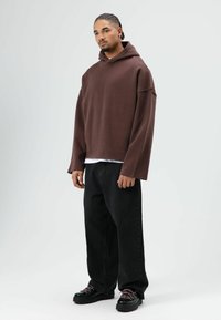 Sweat à capuche marron oversize, avec capuche et ourlet brut, associé à un jean noir ample et des chaussures noires épaisses avec des lacets roses.