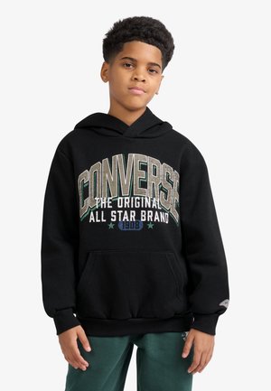 Dreng med kort krøllet hår iført sort Converse hættetrøje med stort hvidt og grønt logo og grønne bukser mod hvid baggrund.