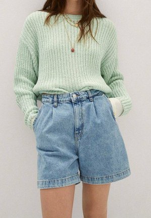 Vrouw draagt een mintgroene gebreide trui en lichtblauwe, hooggetailleerde denim shorts met handen in de zakken.