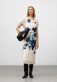 Vrouw in een geplooide witte midi-jurk met blauwe inktvlekpatronen, zwarte kniehoge laarzen, terwijl ze een opgevouwen zwart leren jasje vasthoudt.