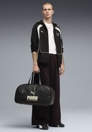 Junger Mann in einem schwarz-weißen Puma-Trainingsanzug hält eine große schwarze Puma-Reisetasche und steht vor einem schlichten grauen Hintergrund.