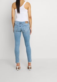 Žena v modrých úzkých džínách, bílém topu přes jedno rameno a bílých vysokých podpatcích stojící na jednobarevném pozadí.