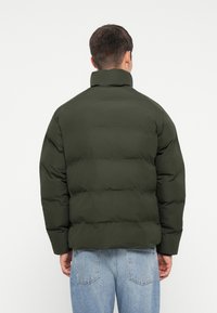 Paljāks žakets tumši zaļā krāsā, ar augstu apkakli, quilted tekstūru un kvadrātveida siluetu, nēsāts ar gaiši zilajiem džinsiem.