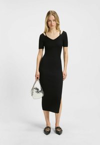 Robe midi noire côtelée à manches courtes, avec un décolleté en V et une fente sur le côté. Associée à un sac transparent en forme de cœur et des ballerines à bout pointu.