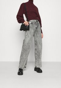 Jean large gris avec une finition délavée, associé à un pull col roulé en tricot bordeaux et un sac bandoulière noir. Des bottes noires à talons larges sont portées.