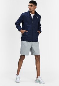 Veste coupe-vent marine avec col montant, fermeture éclair et poignets élastiques. Shorts gris clair et baskets blanches avec des accents pastel.