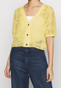 Geel gehaakte cardigan met een V-hals, korte pofmouwen en vier knopen. Heeft een bloemenpatroon en een lichte, ademende textuur.