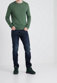 Grüner Strickpullover mit gerippten Bündchen und Saum, kombiniert mit dunkelblauen Jeans und grünen Sneakers mit schwarzen Akzenten und goldfarbenen Ösen.