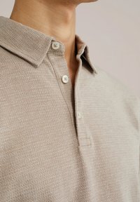 Beige textúrájú pólóing, gallérral, három fehér gombbal, és egy finom mintával, ami mélységet ad az anyagnak.