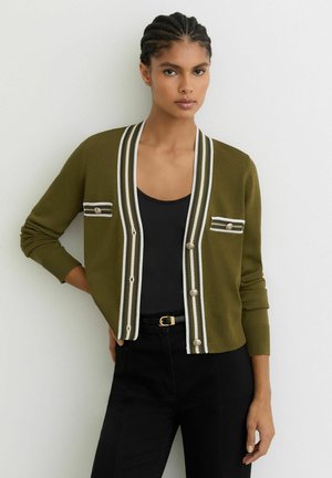 Olíva zöld kardigán V-nyakkivágással, fehér és fekete csíkos szegéllyel, két gombos első zsebbel és bordázott mandzsettákkal, fekete felső fölött viselve.