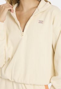 Πουλόβερ σε εκρού χρώμα με φερμουάρ τριών τετάρτων, ριμπ ύφανση και κεντημένο λογότυπο. Διαθέτει μακριά μανίκια και ελαστική λεπτομέρεια στη μέση.