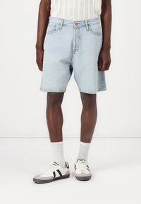 Shorts de mezclilla azul claro con un corte relajado, cinco bolsillos, presillas para cinturón y cierre de botón. Combinados con calcetines blancos y zapatillas con detalles negros.