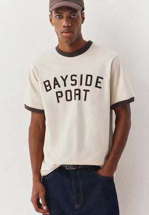 Mladi moški, oblečen v bež in rjavo majico s napisom "BAYSIDE PORT", temno modre kavbojke in rjavo kapo, stoji ob običajni beli steni.