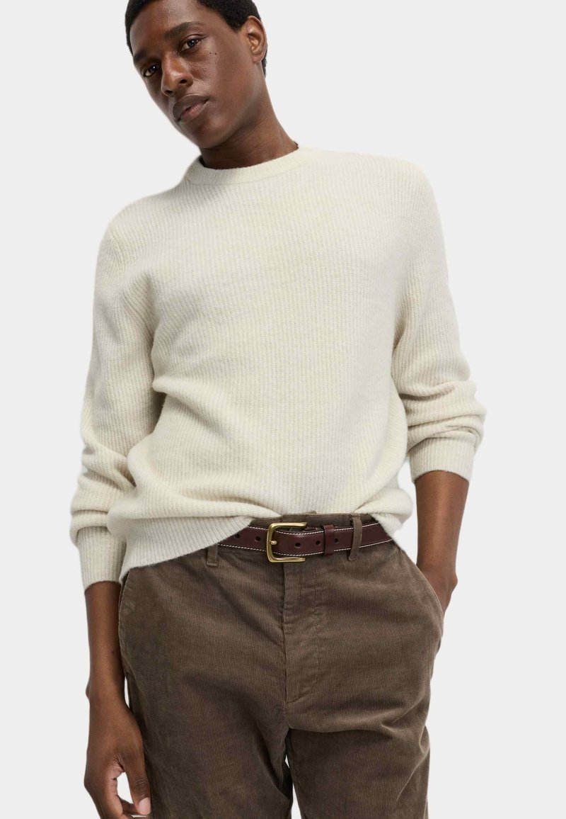 Cremefarbener, ribbeliger Strickpullover mit langen Ärmeln, kombiniert mit braunen Cordhosen und einem braunen Ledergürtel mit goldener Schnalle.