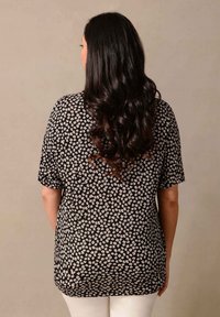 Černý top s květinovým vzorem v bílé a světle růžové barvě. Má krátké rukávy a mírně delší zadní lem. Hladký textilní povrch.