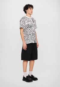 Grafiska t-kreklu ar melnu un baltu tekstu, saskaņota ar melnām šortiem. Modelis valkā melnas slaidējošās kurpes un baltas zeķes.