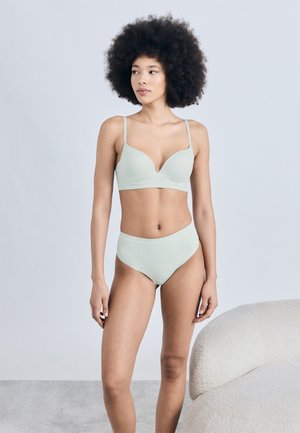 Γυναίκα με μεγάλα μπούκλα μαλλιά που φοράει ανοιχτό πράσινο σουτιέν και εσώρουχο με ψηλή μέση, στέκεται δίπλα σε μια υφασμάτινη λευκή πολυθρόνα μπροστά σε ένα απλό τοίχο.