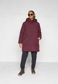 Doudoune bordeaux avec capuche, fermeture éclair à l'avant, deux grandes poches et une doublure intérieure contrastante. Portée avec un pantalon foncé et des chaussures marron.