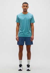 Άνδρας όρθιος, φοράει τυρκουάζ αθλητική μπλούζα, σκούρο μπλε σορτς, λευκές κάλτσες και λευκά παπούτσια τρεξίματος μπροστά σε απλό λευκό φόντο.