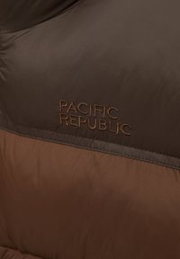 Brun puffjacka med "PACIFIC REPUBLIC" broderat i mörkbrunt på framsidan, som visar en tvåfärgad design med varierande nyanser.