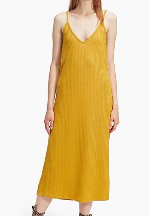 Femme portant une robe midi sans manches moutarde côtelée avec de fines bretelles et un décolleté en V, debout, les bras détendus le long du corps.
