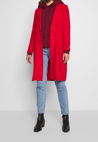 Rode jas met een lange snit, gemaakt van een zachte stof, met een kraag en zijzakken, gedragen over een bordeauxkleurige hoodie en blauwe jeans.