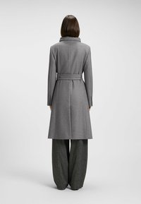 Cappotto di lana grigio con un colletto strutturato, vita cintrata e lunghezza al di sotto del ginocchio. Tessuto strutturato con una finitura liscia, abbinato a pantaloni ampi grigi.