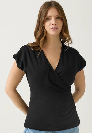 Frau mit schulterlangem, welligem Haar, die ein schwarzes, kurzärmeliges Wickeloberteil und hellblaue Jeans trägt und vor einem schlichten Hintergrund steht.