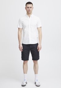 Witte kortearmige overhemdje met knopen, gecombineerd met zwarte cargoshorts met zijzakken en een relaxed fit, gedragen met witte en grijze sneakers.