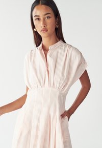 Vestido a rayas en rosa claro y blanco, con mangas cortas, tapeta de botones, cintura ceñida y bolsillos laterales. Textura de tela suave.
