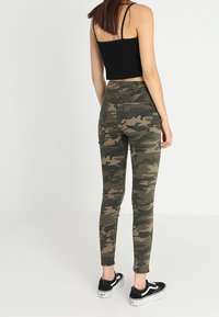 Kamouflage skinny jeans i olivgrön med hög midja, fram- och bakfickor, samt ankellång skärning, kombinerat med svarta sneakers.