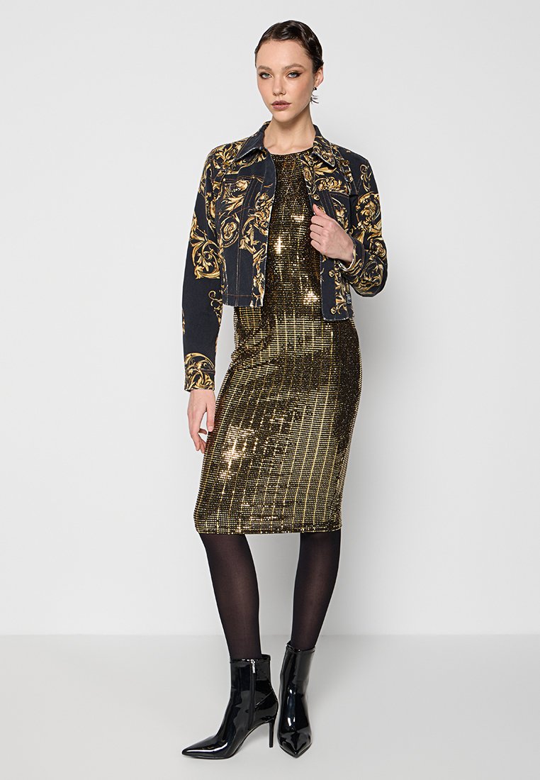 Chaqueta de mezclilla oscura con un patrón floral dorado, combinada con un vestido ajustado con lentejuelas doradas y botines negros, creando un look en capas.