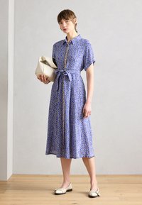 Vestido azul com padrão, com frente abotoada, mangas curtas e cintura atada. O modelo segura uma bolsa de cor bege enquanto usa sapatos rasos.