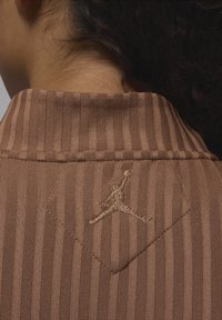 Brunstripete stoffkrage med et diamantformet merke som har en gullbrodert Jumpman-logo. Teksturen ser glatt og fint vevd ut.