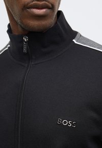 Čierna bunda na zips s vysokým golierom, šedými akcentmi na ramenách a kovovým logom „BOSS“ na hrudi. Textúrovaná látka s hladkým povrchom.