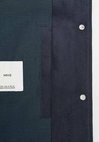 Tmavá látková bunda kombinující různé textury s hladkým povrchem a semišovým vzhledem. Obsahuje bílou štítek s logem a pokyny pro péči.