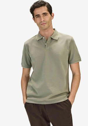 Mann mit dunklen Haaren, der ein hellgrünes kurzärmeliges Poloshirt und eine dunkle Hose trägt, mit den Händen in den Taschen, vor einem schlichten weißen Hintergrund.