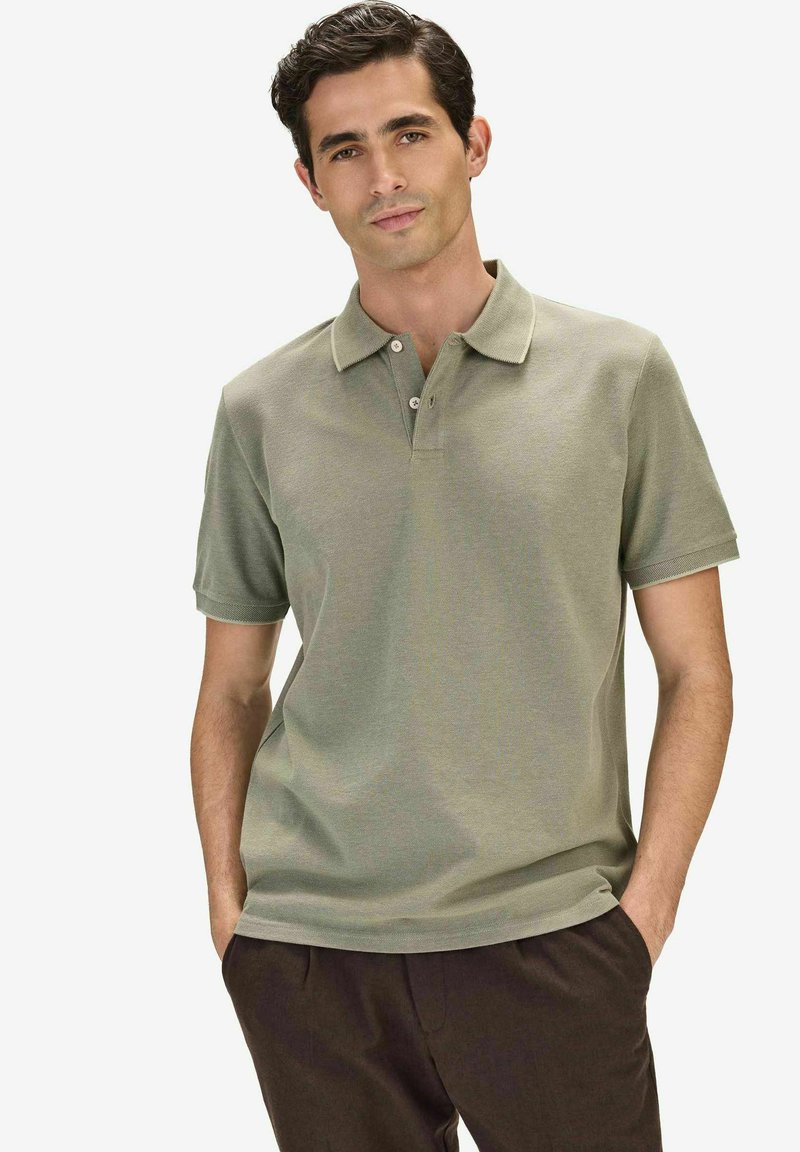 Homme aux cheveux foncés portant un polo à manches courtes vert clair et un pantalon foncé, les mains dans les poches, sur un fond blanc uni.