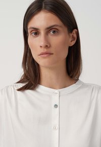 Indumento bianco con colletto a banda, caratterizzato da una placca con due bottoni; tessuto morbido con un motivo sottile.