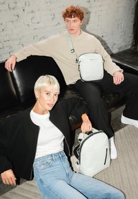 Rucksack in Hellgrau, aus weichem Material mit schwarzen Akzenten und Reißverschluss. Modell in schwarzer Bomberjacke und Jeans, sitzt auf einem dunklen Sofa.
