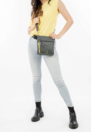 Borsa a tracolla grigia in tessuto testurizzato, con chiusura a zip gialla e tracolla regolabile; tenuta da una persona che indossa jeans azzurri e stivali neri.